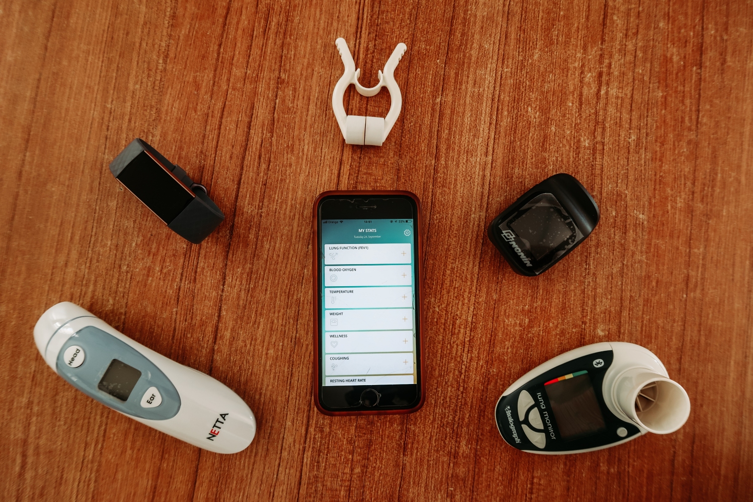 Medical equipment, including a thermometer, smart phone, smart watch and lung function monitor laid out on a brown surface.