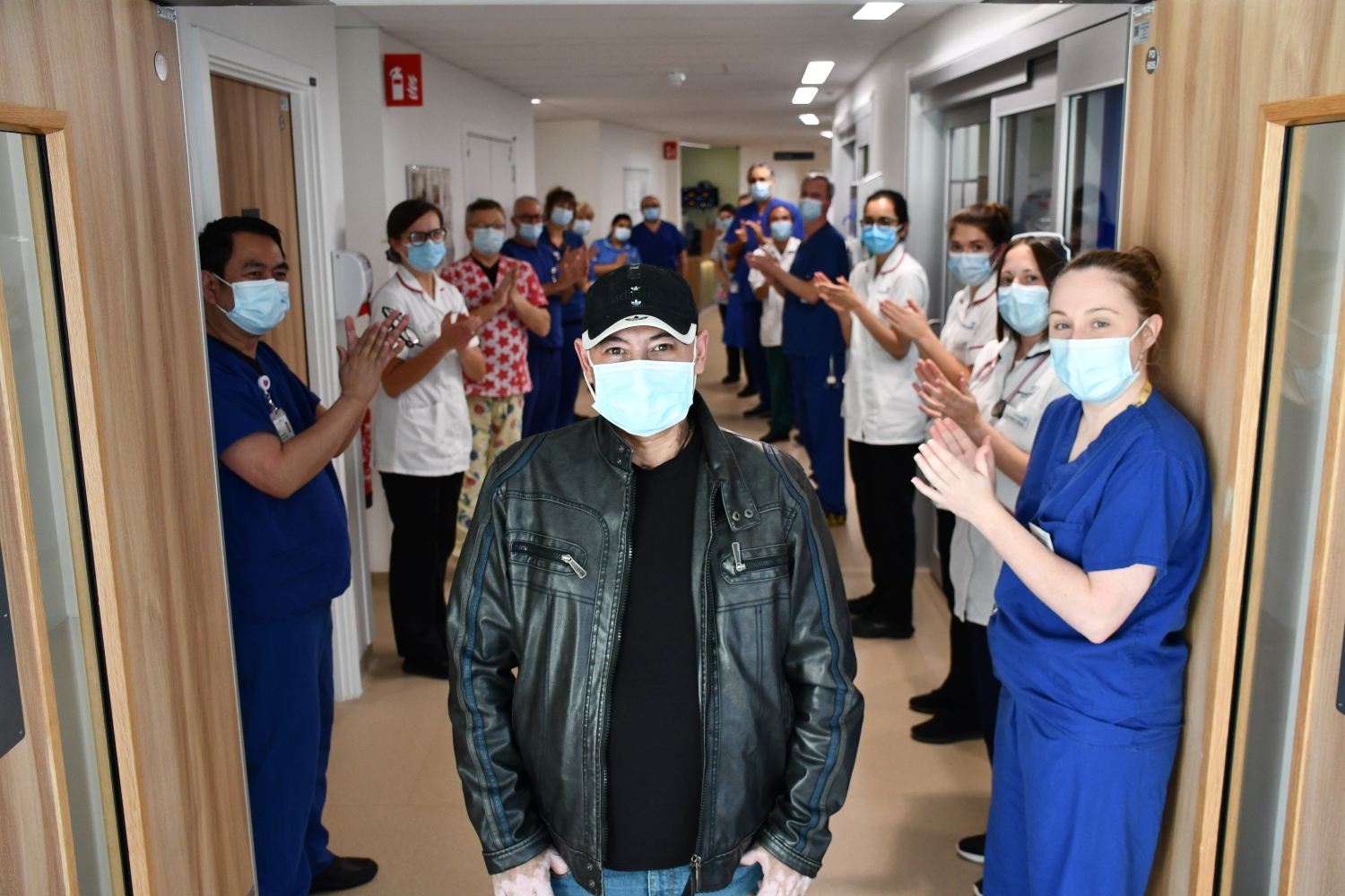 A man wearing a baseball cap, leather jacket and mask stands in a corridor, while staff behind him clap.