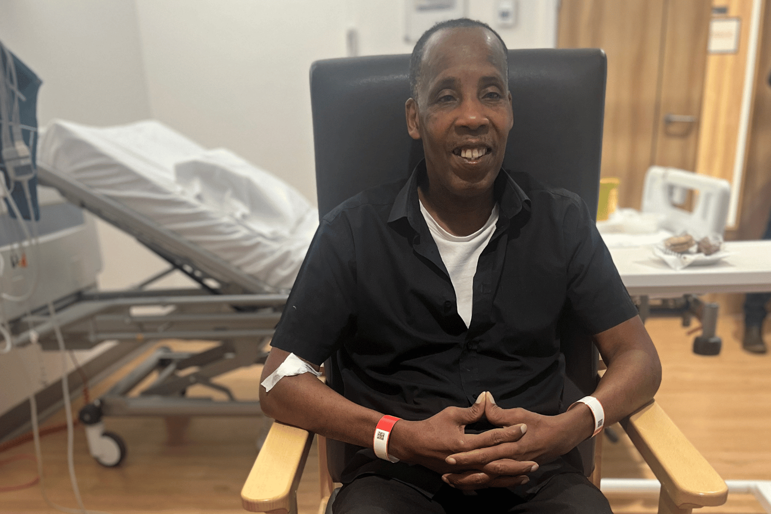 A man sat in a chair in a hospital bedroom, smiling.