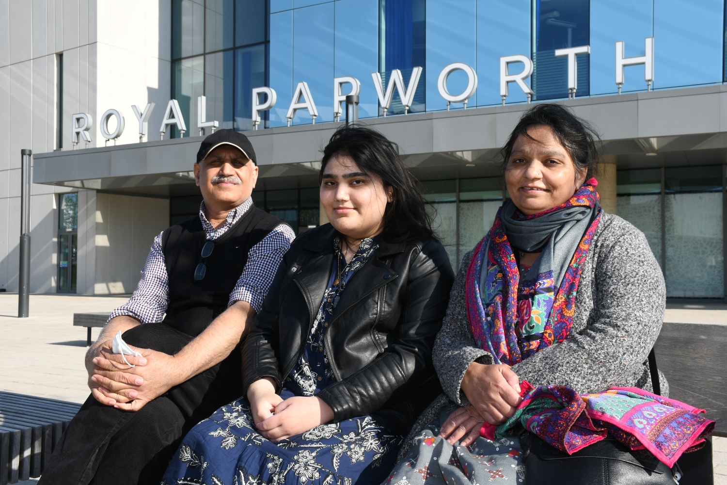Areeb, mum and dad sitting down outside a blue building with the letters 'Royal Papworth'