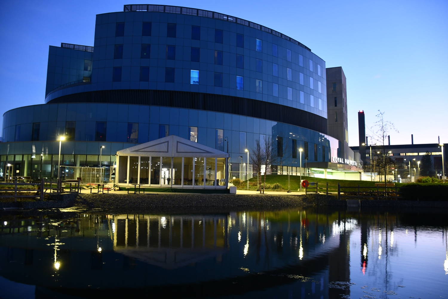 A blue oval shaped building, with a pond in the foreground, in early morning dusk. 