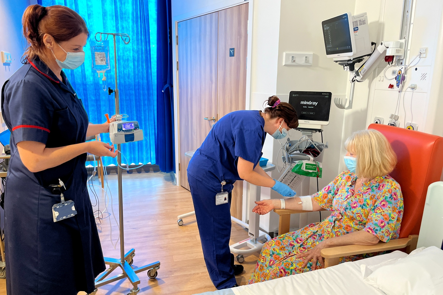 A nurse stands next to a drip stand while another healthcare worker in blue scrubs puts a needle into a patient's arm.