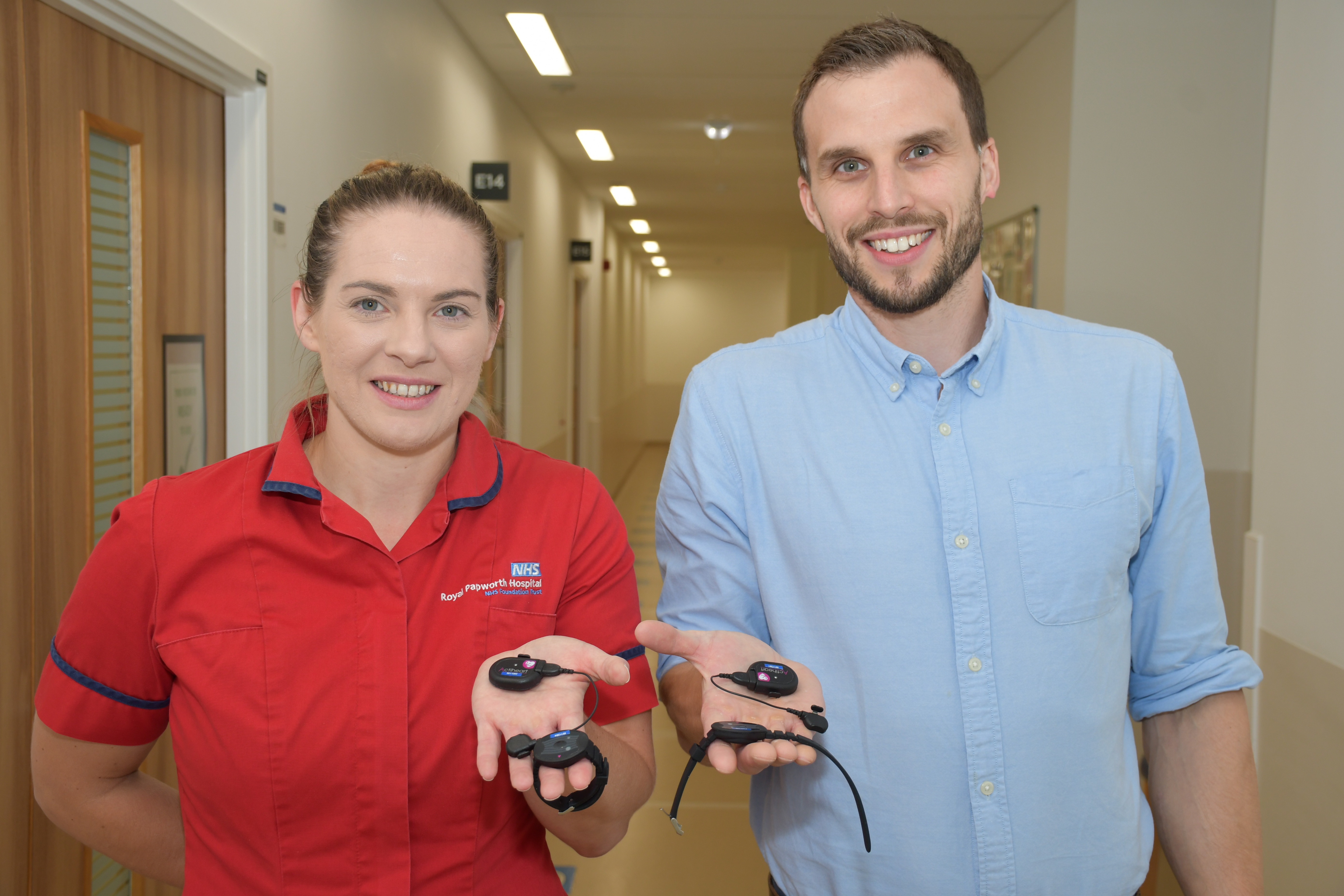 A photo of a woman and a man smiling. The woman is wearing a red healthcare tunic and the man is wearing a blue shirt. They are both holding black wearable technology devices. 