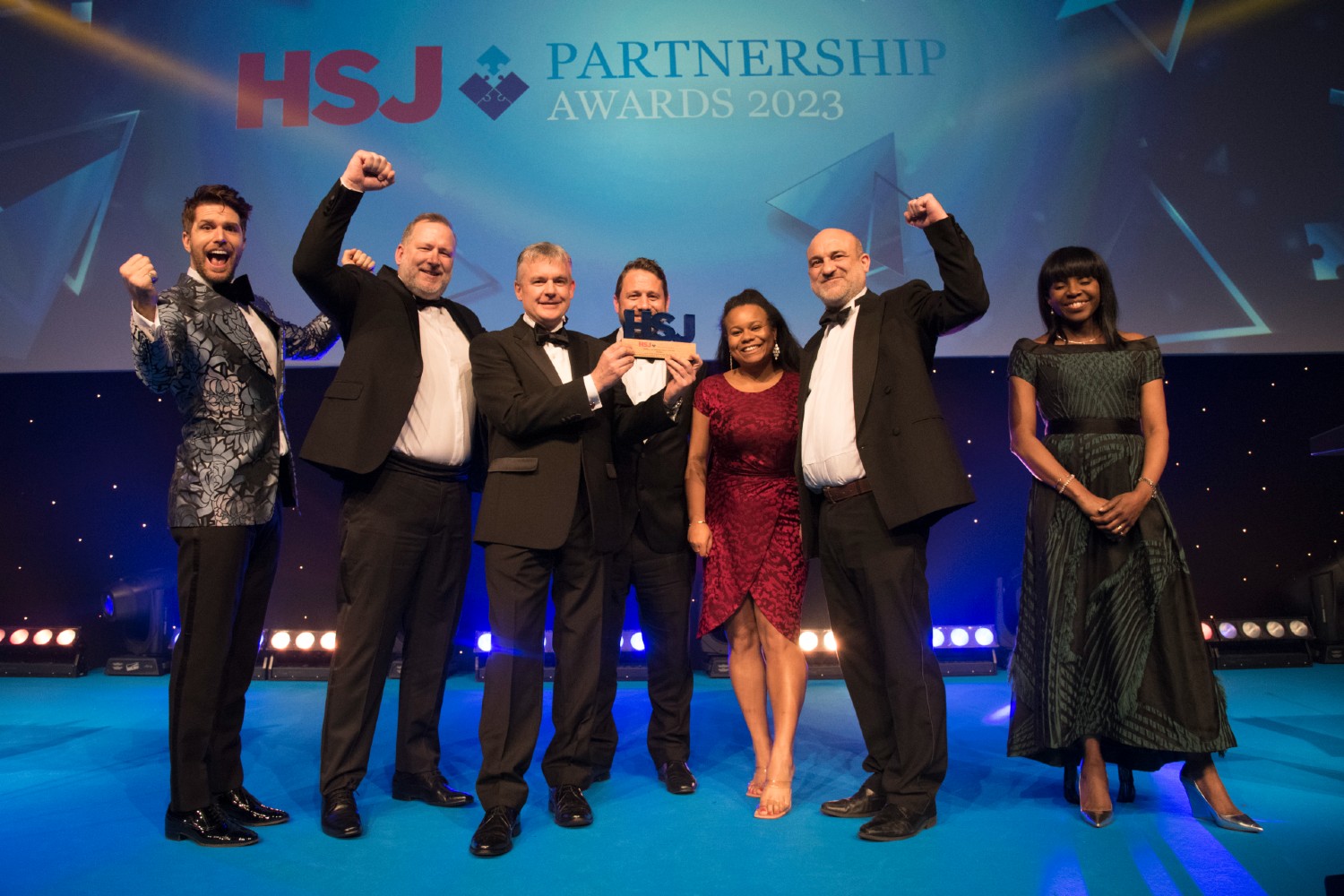 Seven people on stage wearing suits and dresses holding an award in front of a background that says: HSJ Partnership Awards
