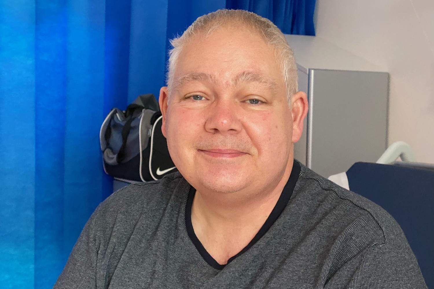 A person in a grey t-shirt, sat in front of a blue curtain