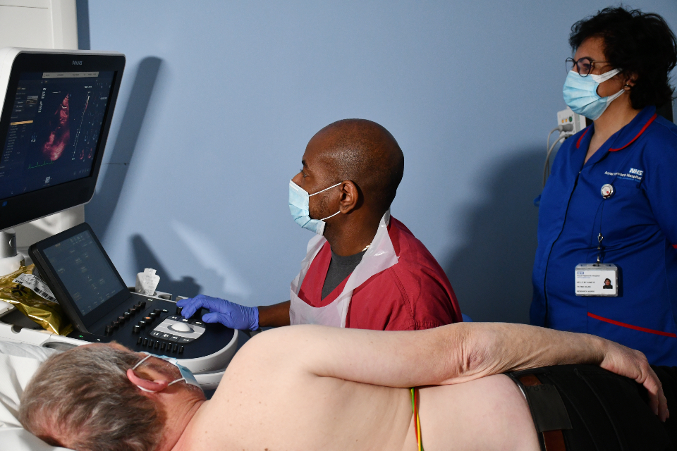 A patient laying on a table, while a person in raspberry coloured scrubs performs an echocardiogram