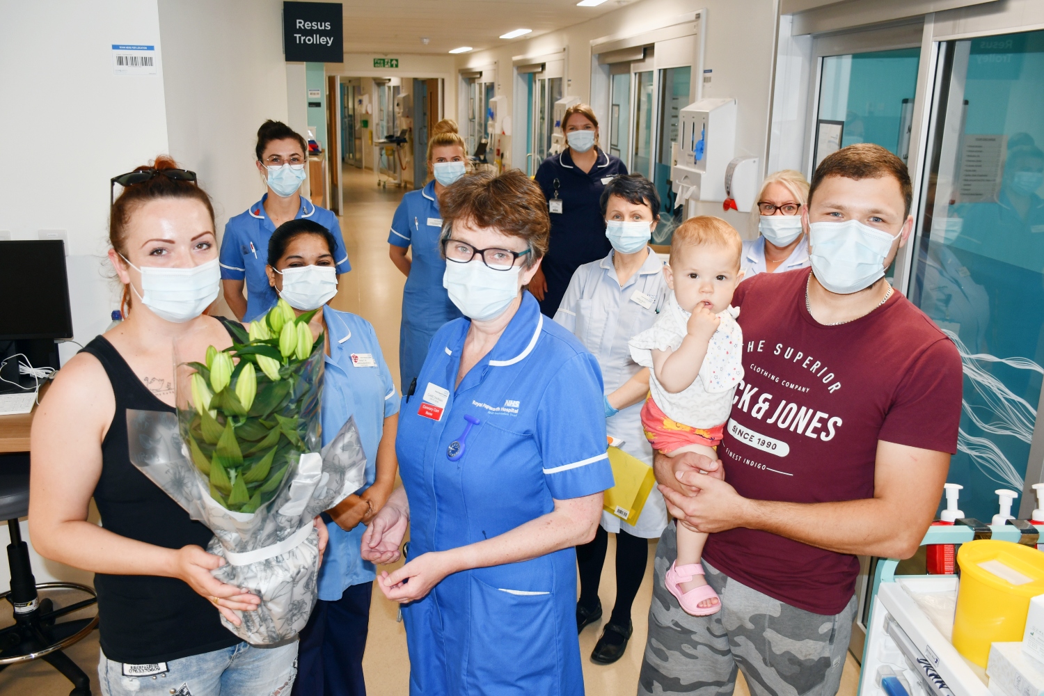 Claire in a blue nurse uniform being presented with a pot of flowers by Monika. Other nurses stand in the background of the ward corridor.