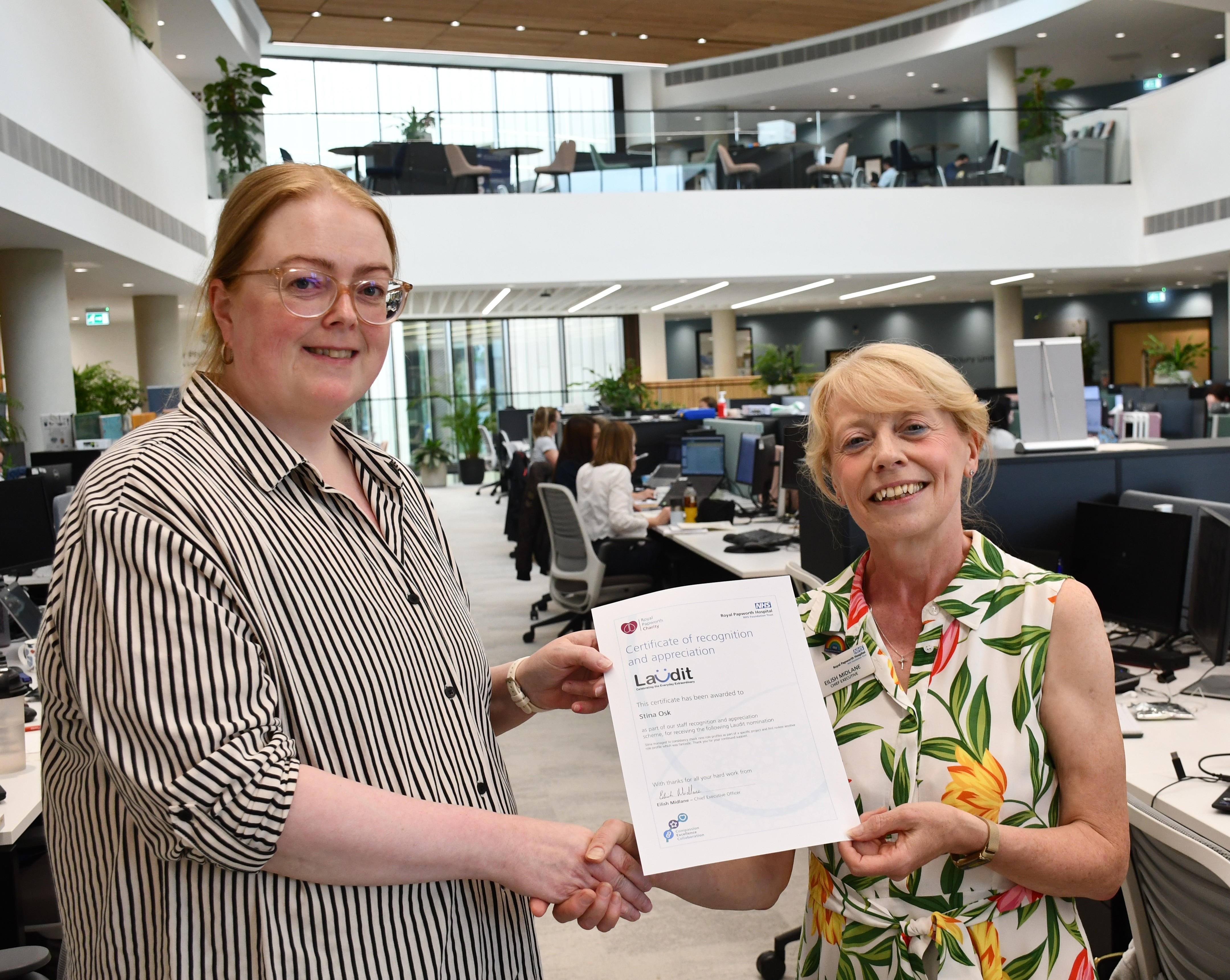 Photo of two women standing in an office, shaking hands and smiling. The lady on the left is wearing a stripy blouse and glasses and being presented with a certificate.