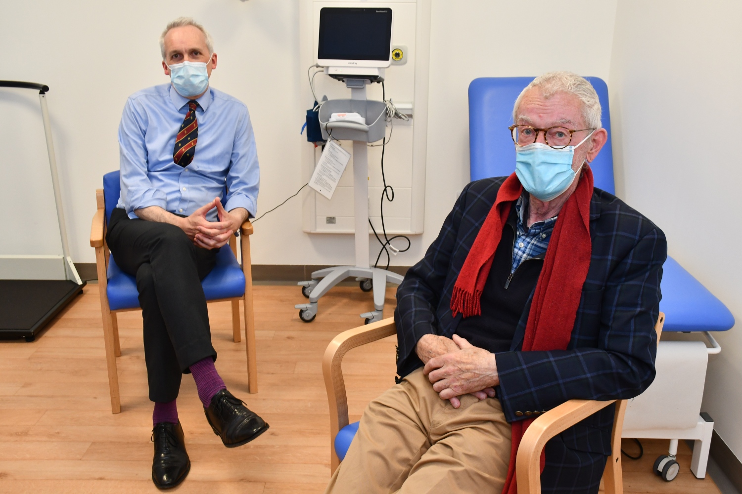 Two people sitting in chairs in a hospital clinic room, wearing masks.