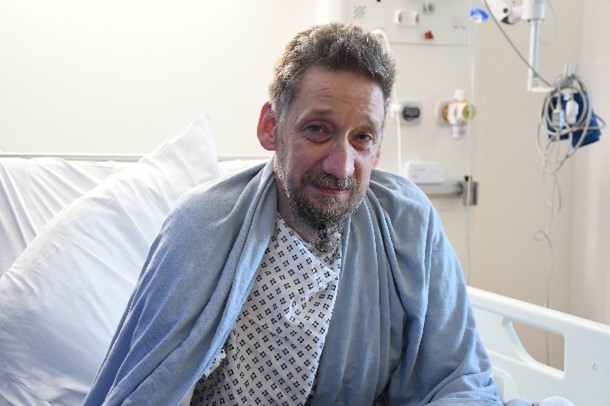 Daniel sitting on a hospital bed, wearing a white and blue pjyama top draped in a light blue throw
