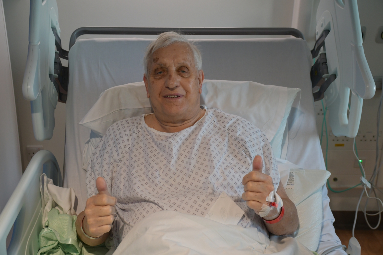 A man sitting up in a hospital bed giving a thumbs up