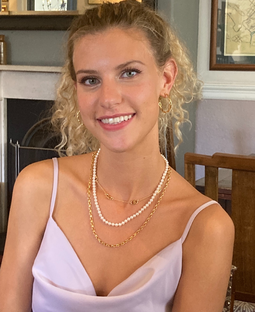 Helena smiling, wearing gold necklaces, pearls and a pink top.