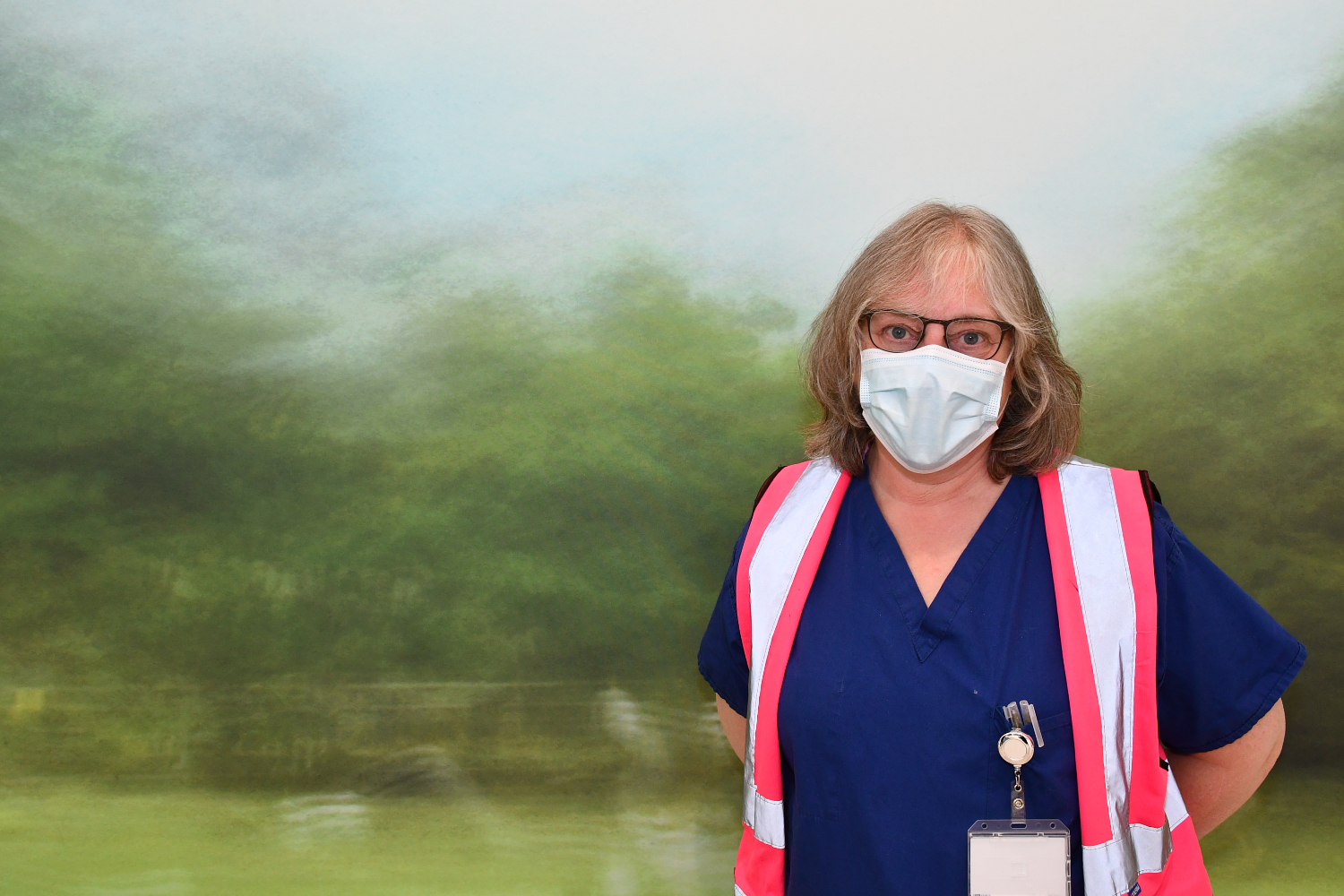 A person wearing glasses and a mask, in blue scrubs with a pink high-vis vest.