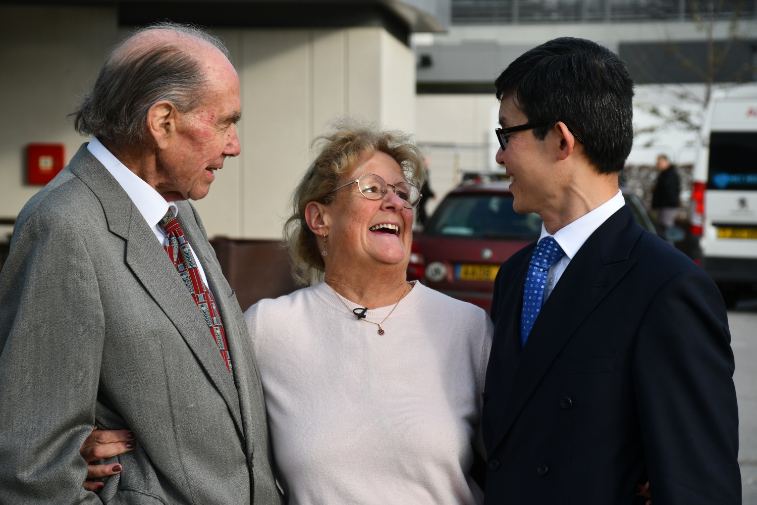 Sandy stands outside between two men in suits, all are smiling and hugging.