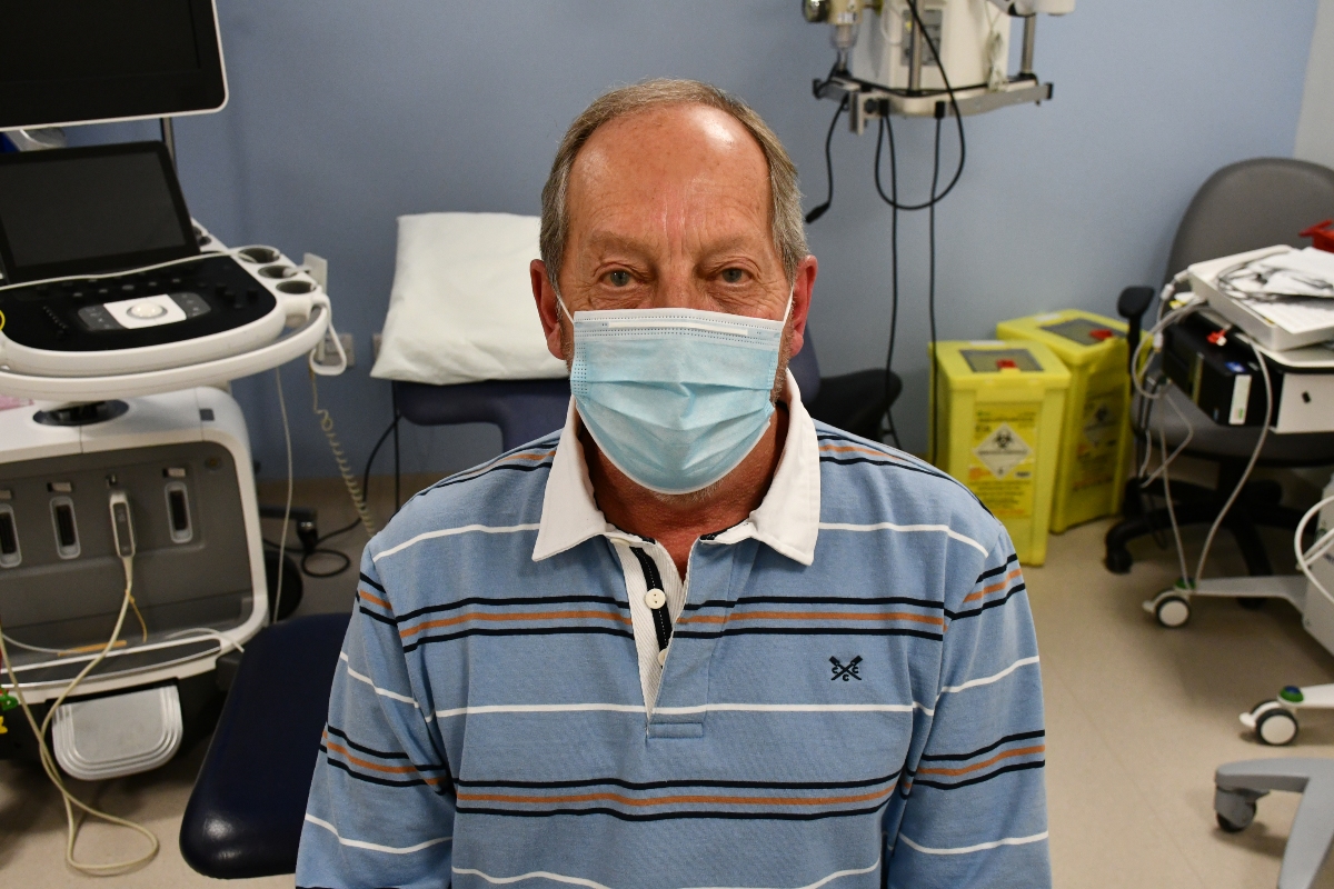A man wearing a mask sitting on a bed, with medical equipment behind him.
