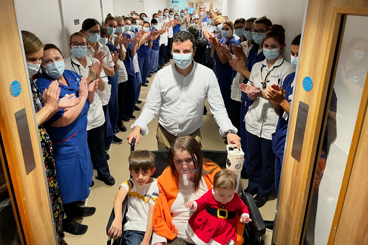 A woman in a wheelchair with her two children. Her husband stands behind them, with NHS staff lining the corridor behind clapping. 