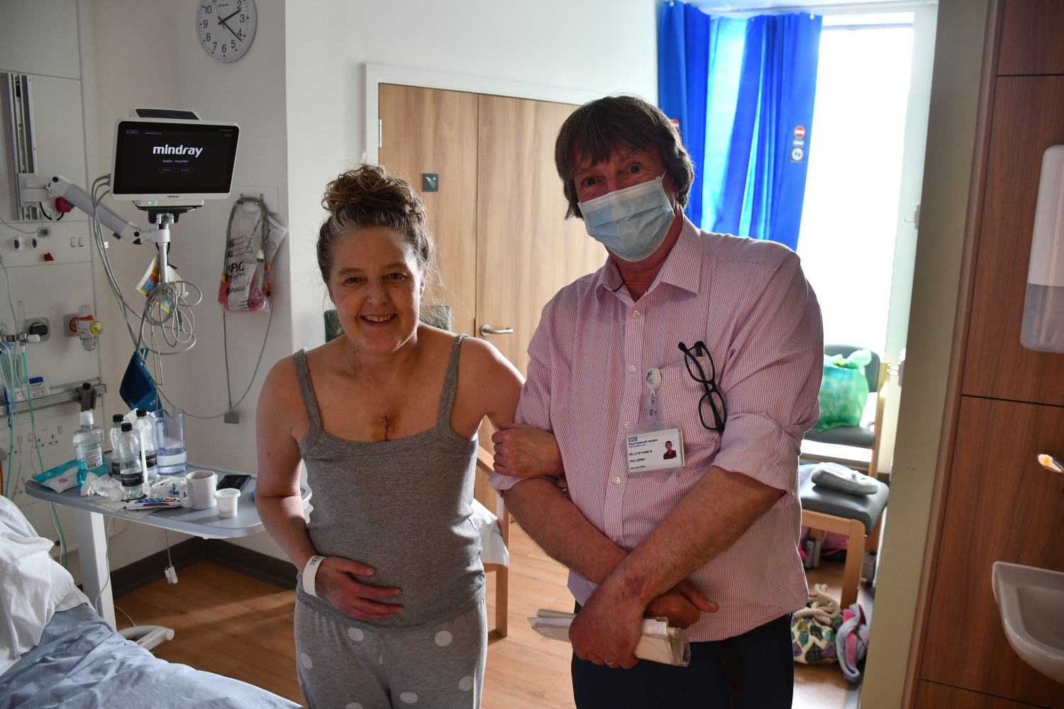 Paul in a pink shirt with a patient wearing a grey vest, holding each others arms and smiling.