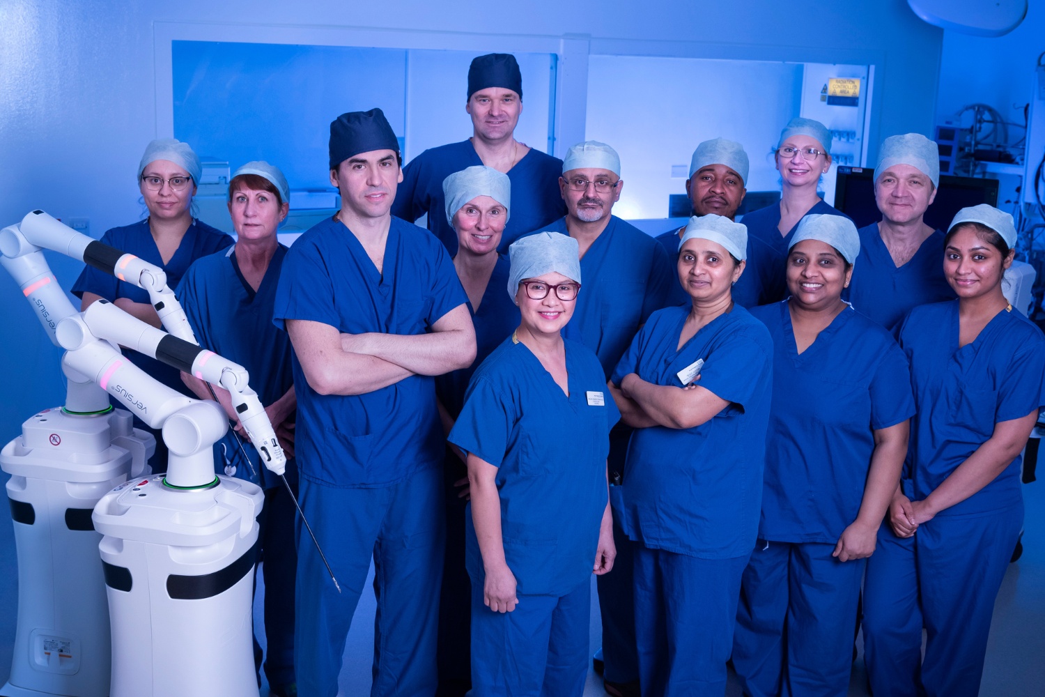 14 people stood in surgical scrubs next to the surgical robot in a surgical theatre.
