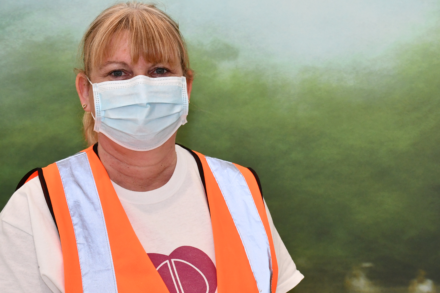 A person with blonde hair, wearing a mask and an orange hi-vis vest.