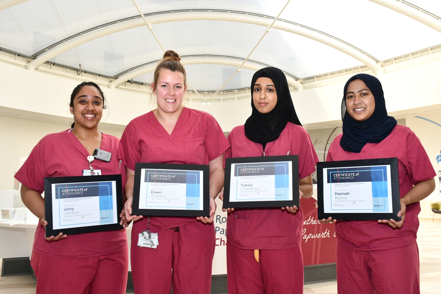 Four people standing indoors, wearing raspberry coloured scrubs, holding certificates and smiling. 