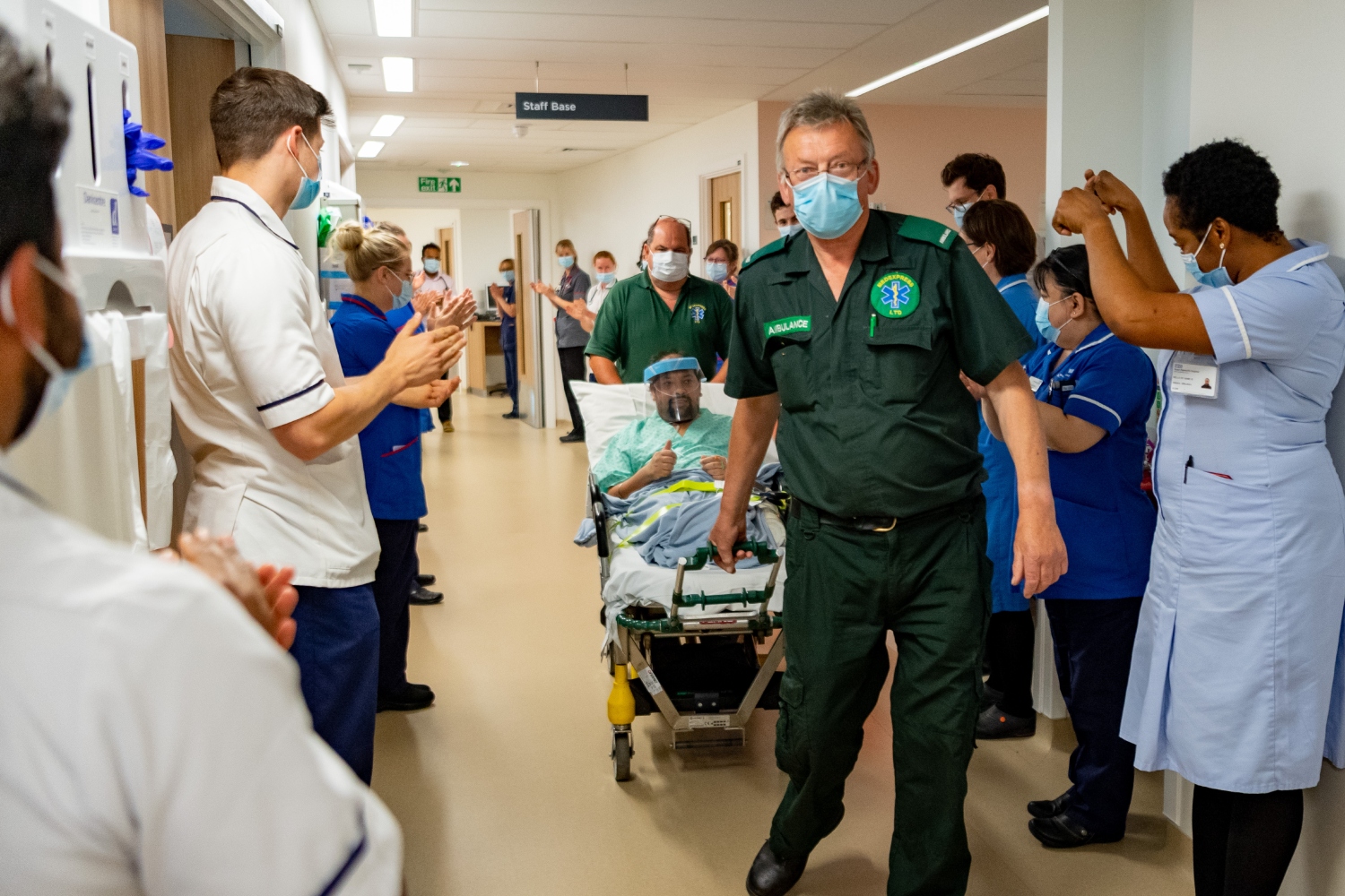 A patient is wheeled down a corridor, with hospital staff clapping either side of the corridor.