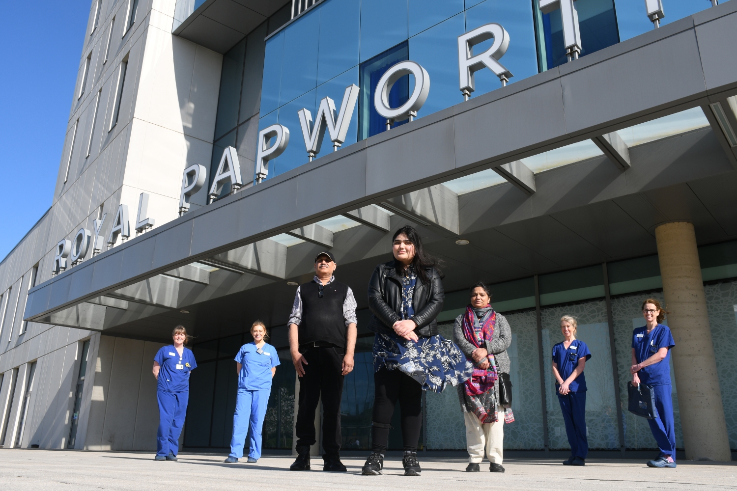 Areeb standing outside a sign saying 'Royal Papworth' with her mum, dad and nurses in blue scrubs either side of her