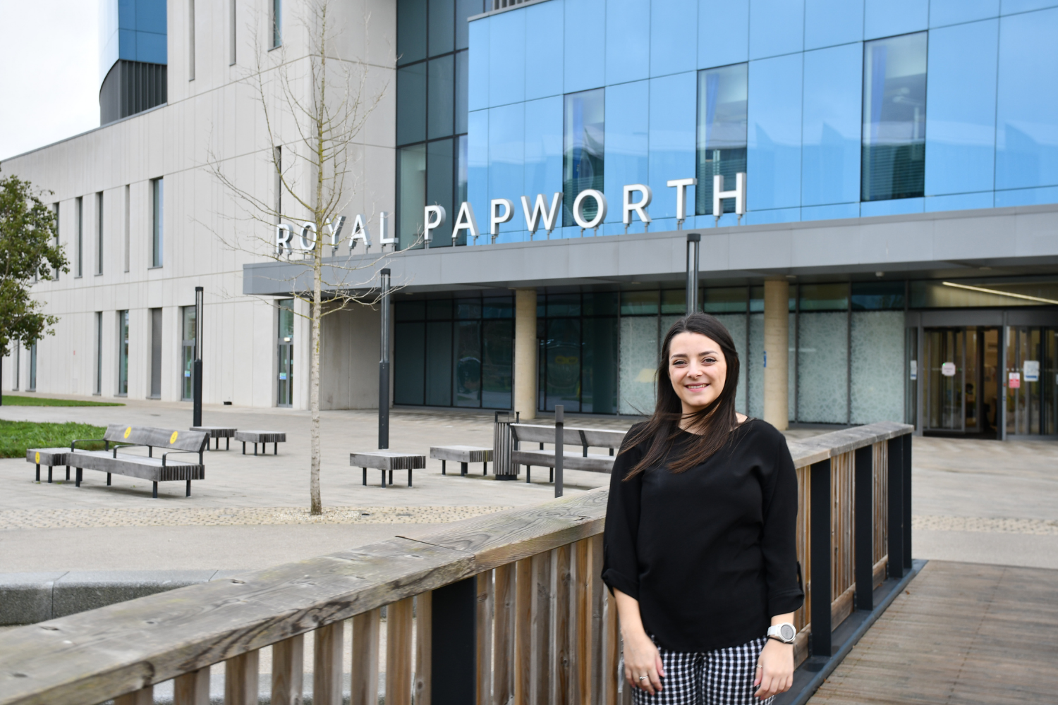 Sustainability officer, Hannah Greensill, standing outside the front entrance to Royal Papworth Hospital, leaning against the bridge and smiling.
