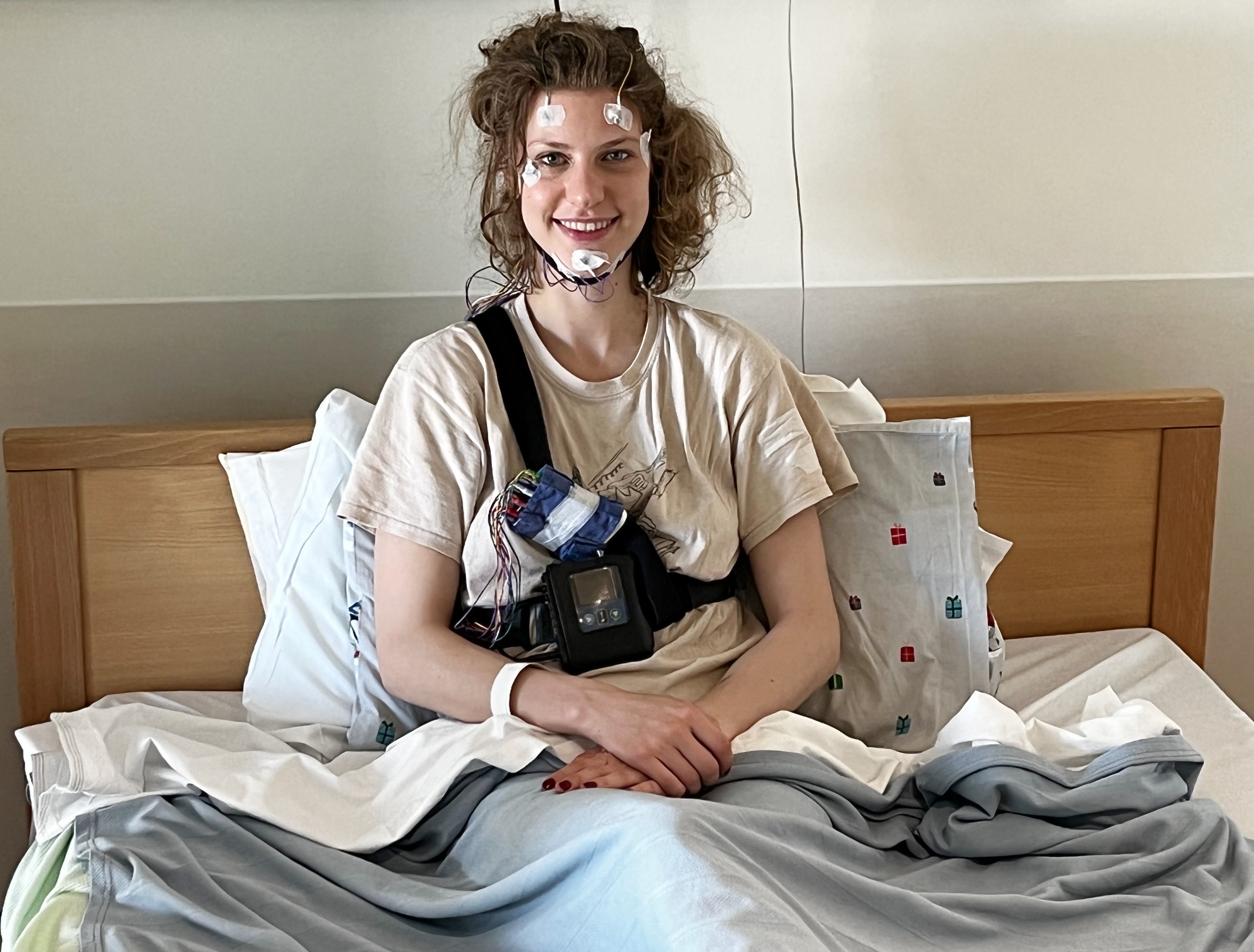 Helena sitting up in a hospital bed, with wires attached to her head and face.