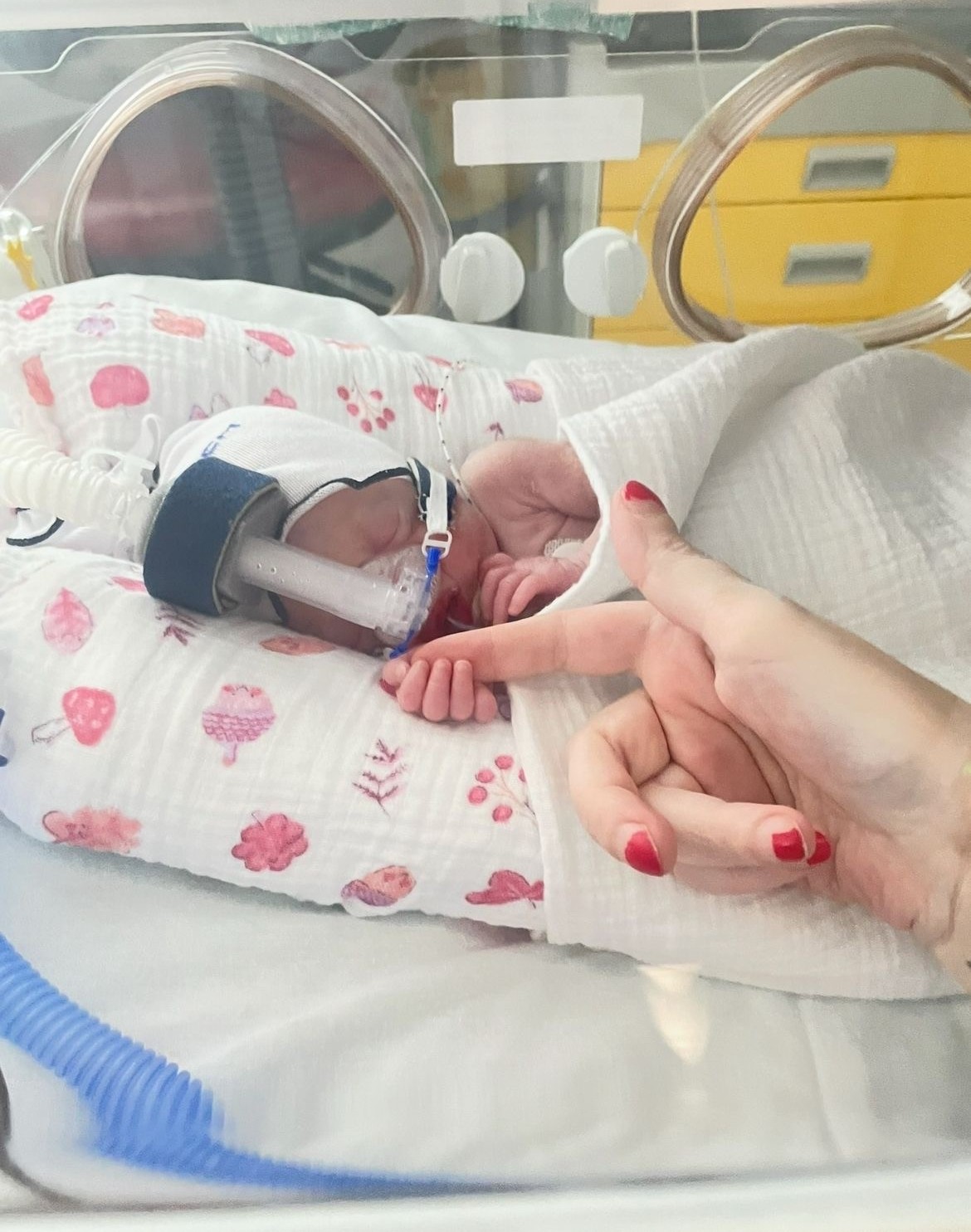 Photo of a tiny baby in an incubator. Its fingers are wrapped around its mother’s index finger.