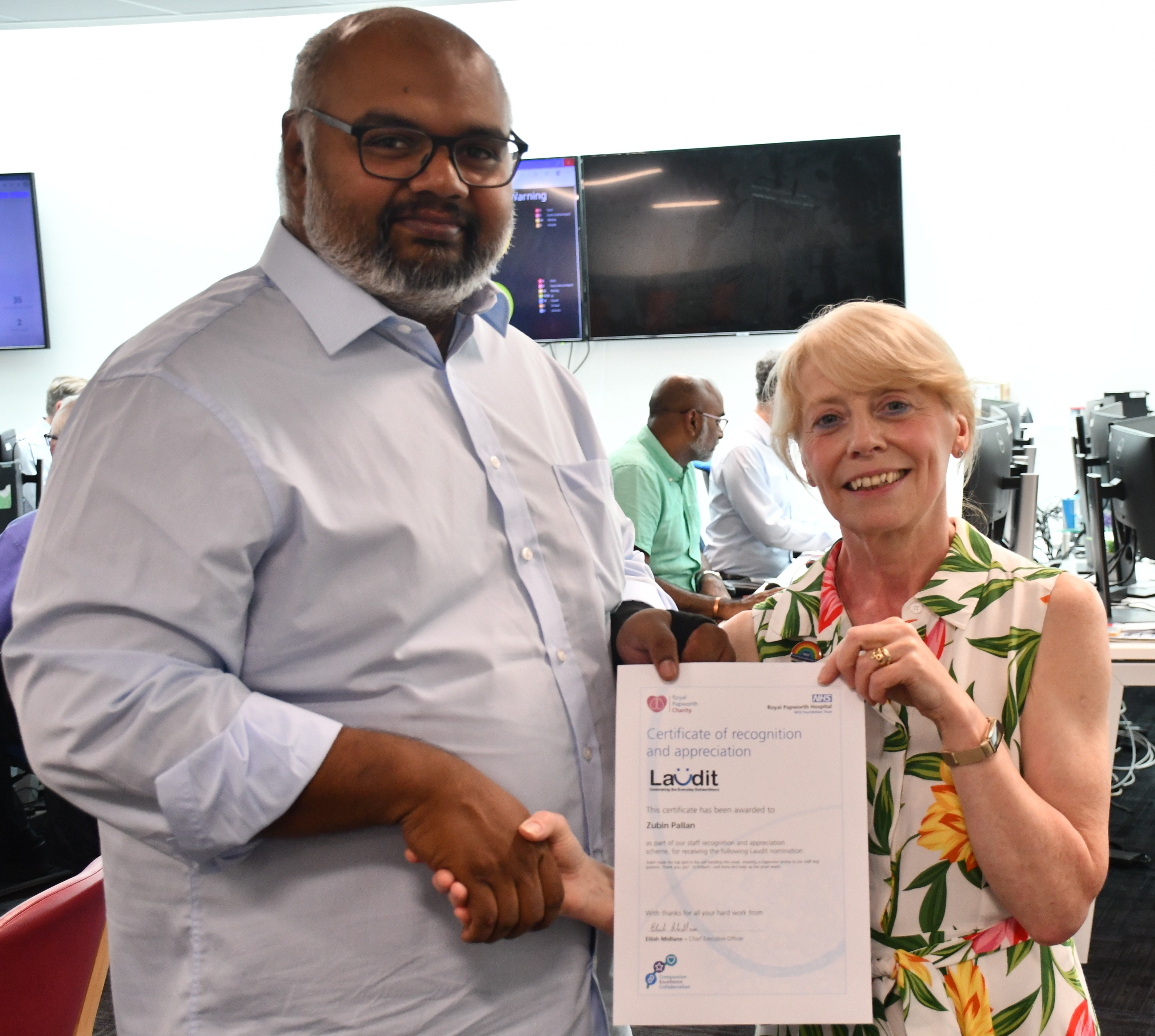 Photo of a man and a woman, shaking hands and smiling.  The man on the left is wearing a blue shirt and glasses and being presented with a certificate. 