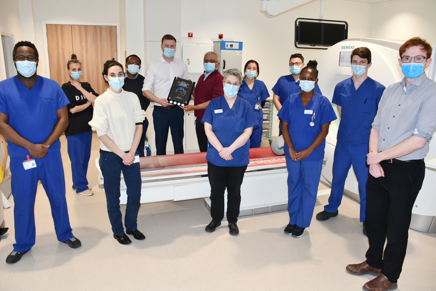 People standing in a CT scan room, with two people in the middle of the room holding a plaque.