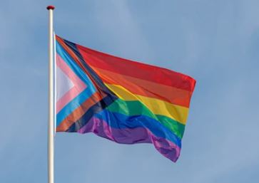 A rainbow flag with red, orange, yellow, green, blue and purple, with an arrow of black, brown, blue, pink and white. 