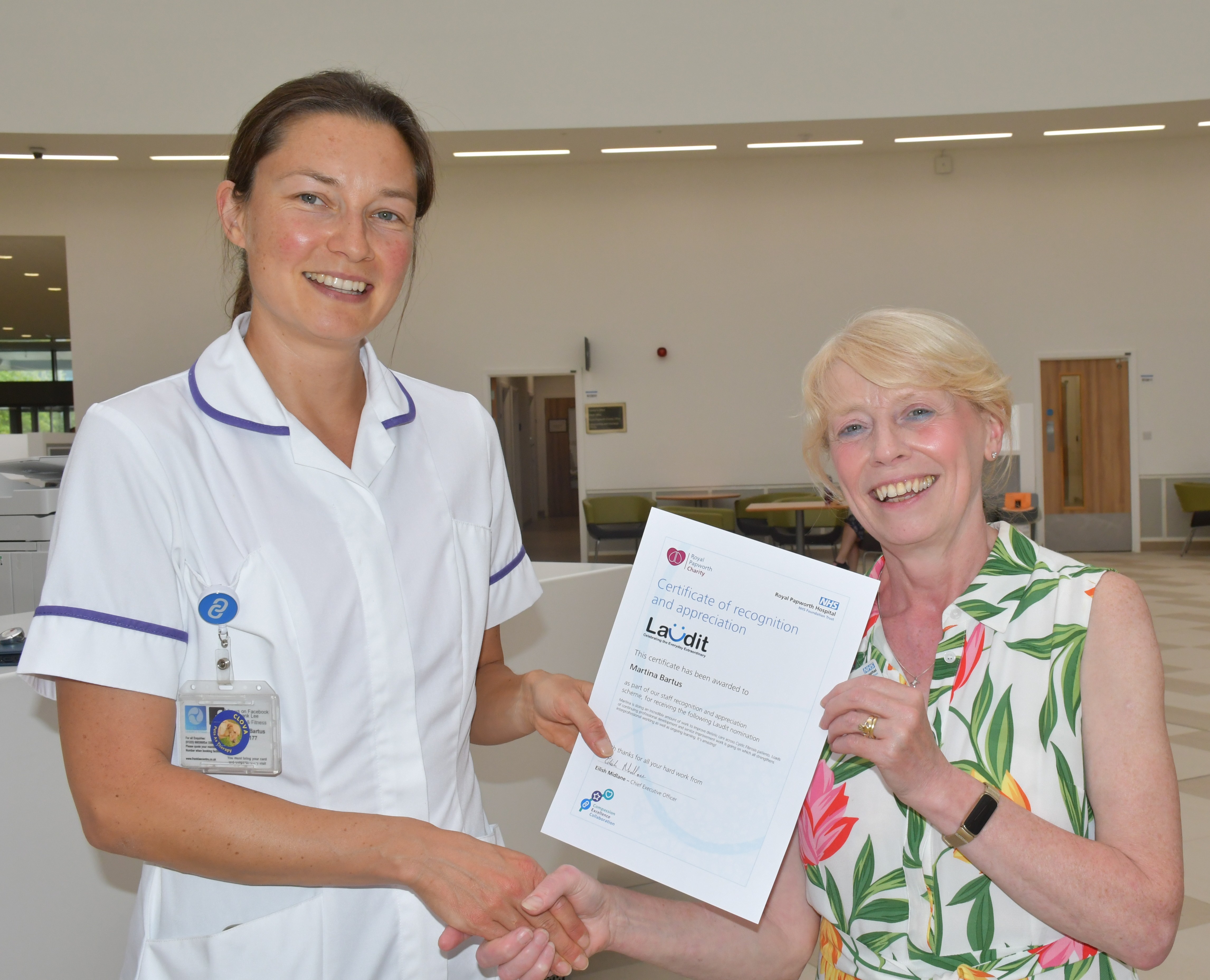Photo of two women standing in a hospital atrium, shaking hands and smiling. The lady on the left is wearing a white tunic and being presented with a certificate.