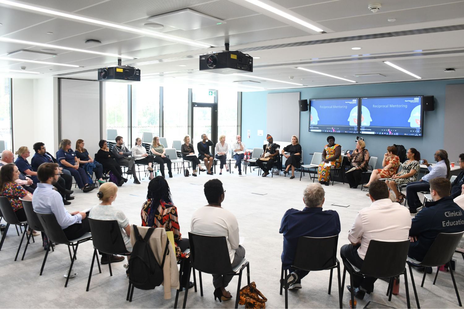 A group of 25 people sitting in a room in a big circle. A screen says 'Reciprocal Mentoring'.